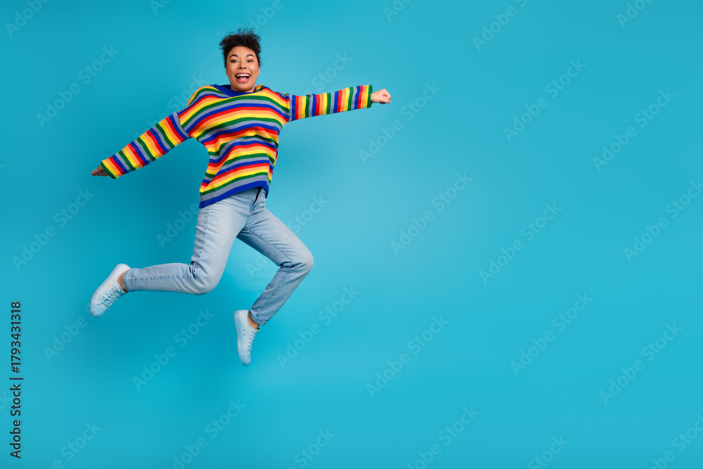 Obraz premium Joyful young woman jumps in striped sweater light jeans against a bold blue background for lifestyle fashion and advertising use