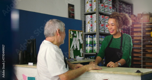 Young woman behind a counter smiling warmly while interacting with an older man in a hardware store with colorful shelves in the background
