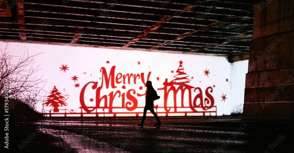 Fototapeta premium Silhouette walks beneath bridge. Red Christmas message visible. Pink light illuminates graffiti, creating stark contrast. Winter scene.