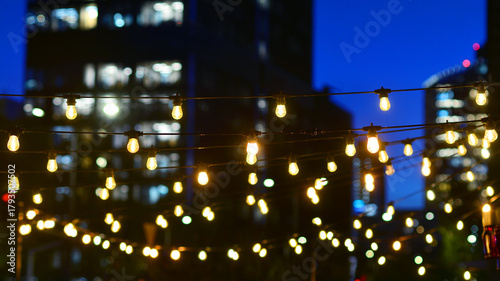 Light bulbs garlands against glass modern buildings background in night. Blurred rows of lit windows out the architectural grid of a modern facade.