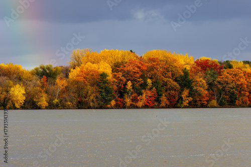 Vibrant autumn foliage lines the shore of a lake, showcasing bright yellow, orange, and red trees under a dramatic sky with a faint rainbow in the distance.