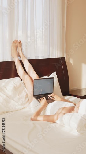 A woman relaxes in bed while working on a laptop, with her legs up and a towel on her head.