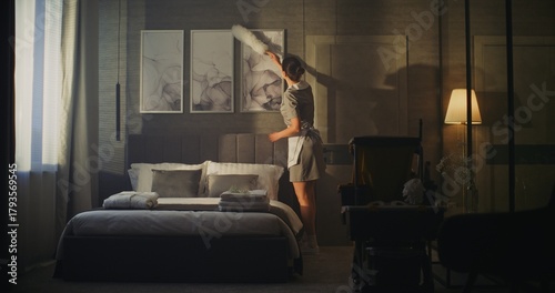 Tableau sur toile Maid in Professional Uniform Gently Cleans Picture Frames on Hotel Room Wall, Removing Dust With White Feather Duster