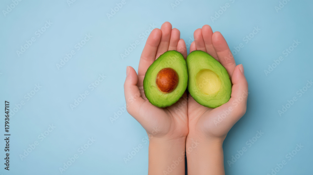 Fototapeta premium Fresh green avocado cut in half with seed held gently in two hands against soft blue background, symbolizing health and natural nutrition