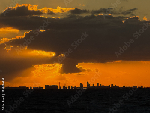 Golden clouds cast glow over city bay, serene ocean horizon captured during vibrant sunset moment