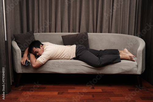 Photos Southeast Asian man in casual clothes lying face down on a grey sofa, falling asleep, appearing exhausted or overwhelmed