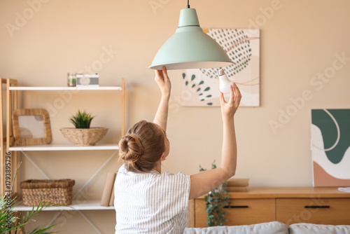 Young woman screwing light bulb into ceiling lamp at home, back view