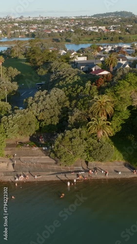 Aerial view of people swimming and relaxing at a beach in Point Chevalier, Auckland, New Zealand. The city skyline is visible in the background. Trees and parkland surround the beach.