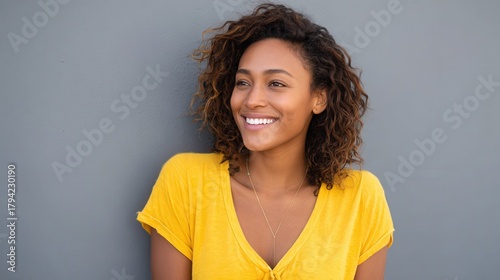 A cheerful woman with curly hair wearing a yellow shirt smiles against a gray wall, conveying happiness and positivity.