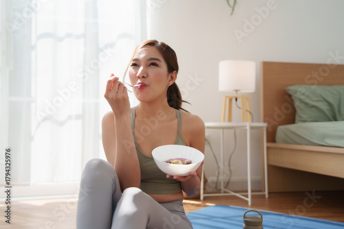 Asian woman in yoga outfit sitting on mat, happily eating fruit yogurt after workout in bright home morning light.