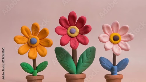 Three colorful clay flowers in pots, yellow, red, and pink, with green and blue leaves, against a light pink background.