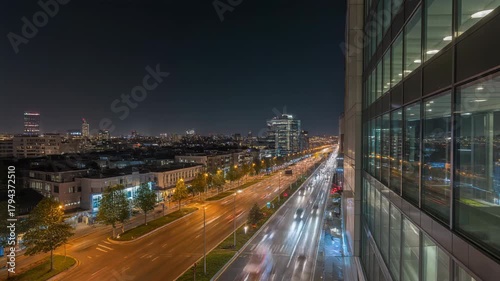 Night view of a city skyline with illuminated buildings and a highway with blurred car lights, seen from a high-rise building.
