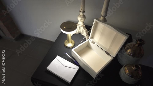 Camera slow spin over hite message box sits open on a black tabletop beside a blue pen and envelope, surrounded by candlesticks, vases, and a lamp, creating a refined and welcoming guest station