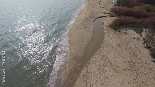 Aerial view of sandy beach with small river flowing into the sea. Coastal landscape with gentle waves, shoreline, and natural water patterns on a cloudy day.
