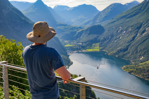Tourist wearing a sun hat enjoys panoramic view of Geiranger Fjord from a scenic lookout in Norway.