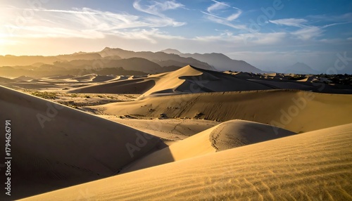 Fototapeta Naklejka Na Ścianę i Meble -  Golden sand dunes roll across a desert landscape under a blue sky, distant mountains silhouetted by sunset