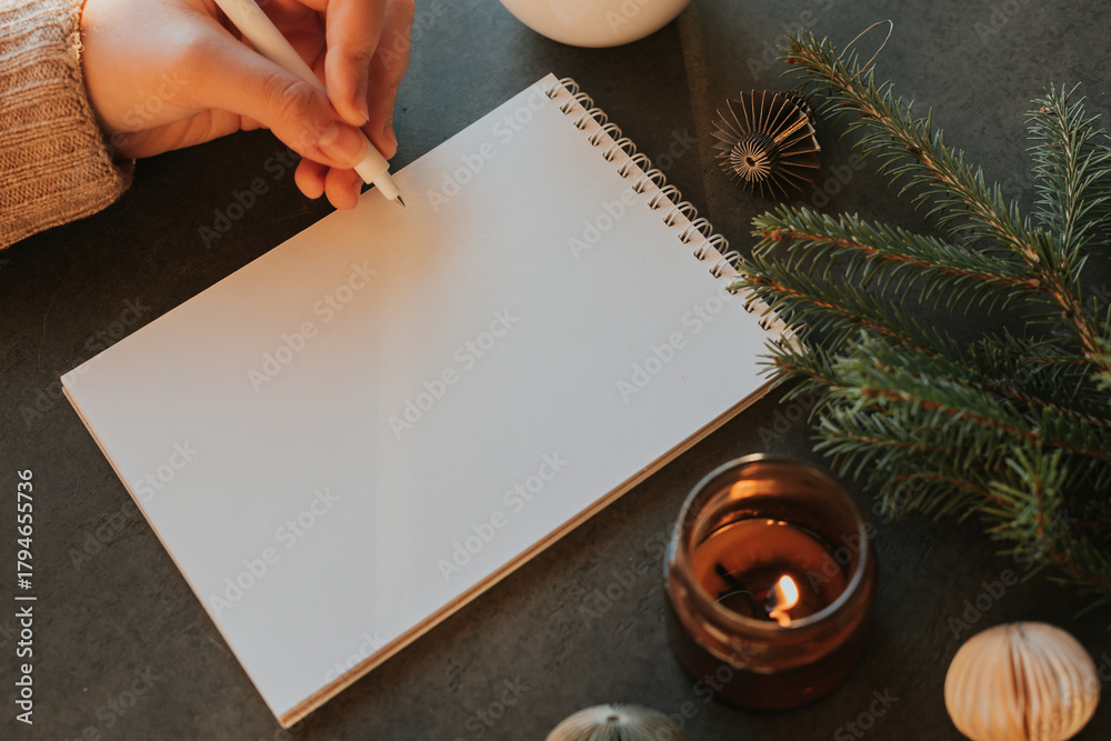 Naklejka premium Female hand writing in notebook. Fir tree, burning candle, cup of coffee and decoration on table. Top view, flat lay. New year's Resolution and Goals