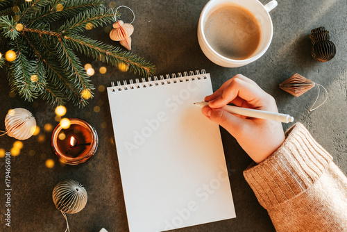 Female hand writing in notebook. Fir tree, burning candle, cup of coffee and decoration on table. Top view, flat lay. New year's Resolution and Goals
