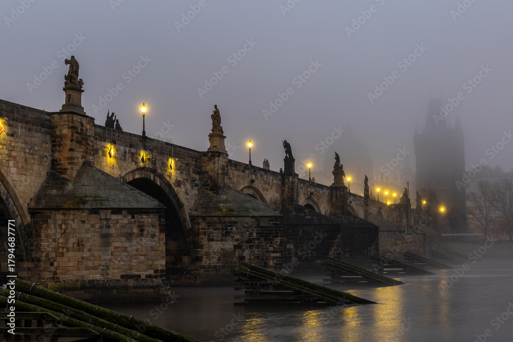 Fototapeta premium Charles Bridge over the Vltava River in Prague, Czech Republic, Shrouded in Morning Fog at Sunrise with Glowing Street Lamps, Empty of People, Creating a Calm and Atmospheric Cityscape