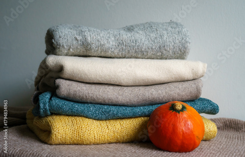 Stack of soft knitted sweaters in neutral tones with a small pumpkin, creating a warm and cozy autumn atmosphere