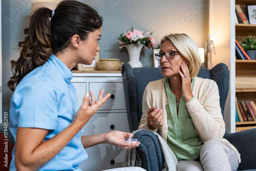 Home hearing care visit: nurse explains and fits a hearing aid for an older woman, gentle coaching and device setup during an in-home audiology consultation to improve communication and daily life