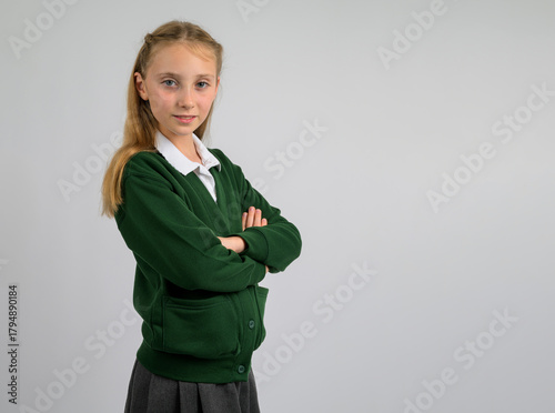 Confident schoolgirl in green cardigan and grey pleated skirt posing with folded arms. Classic British school uniform, children’s autumn fashion and education concept on neutral background