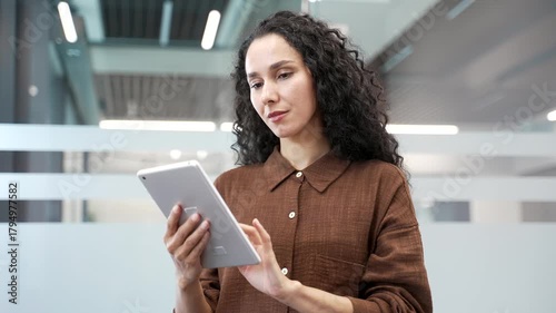 Young serious businesswoman is using digital tablet while standing in a modern business office. Focused beautiful female manager works in application, typing a message or chatting online. Close up