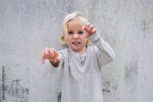 Little 4 Year Old Blonde Girl Standing by a Wall, Pretending to Be Angry and Showing Claws