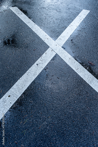 Wet asphalt surface with large white X road marking, crackled paint texture and subtle reflections after rain.
