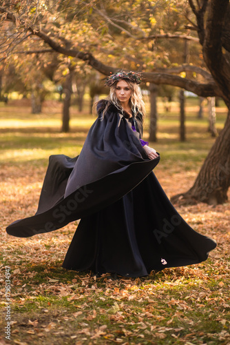 A young woman stands in a golden autumn forest, wearing a black medieval dress with purple accents. She wears a crown of twigs and berries, and a dark hooded cloak. This fantasy portrait  