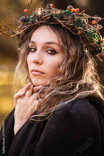 A young woman stands in a golden autumn forest, wearing a black medieval dress with purple accents. She wears a crown of twigs and berries, and a dark hooded cloak. This fantasy portrait  