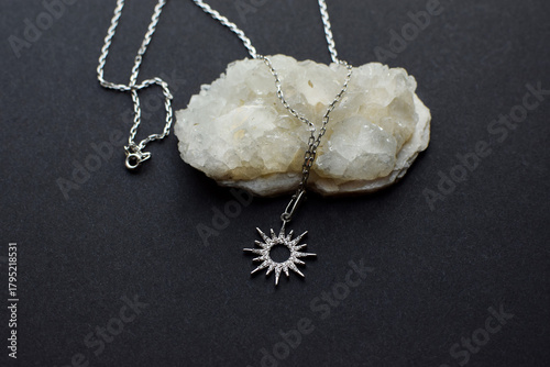 A silver jewelry pendant on a chain in the shape of a sun or a star with stones on a druse with quartz crystals on a dark background.