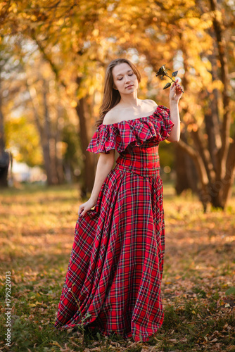 A young woman stands in an autumn forest. She is wearing a long, red, plaid dress with off-the-shoulder details and a frill, a romantic, vintage style. She holds a decorative metal flower in her hands