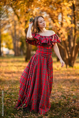 A young woman stands in an autumn forest. She is wearing a long, red, plaid dress with off-the-shoulder details and a frill, a romantic, vintage style. She holds a decorative metal flower in her hands