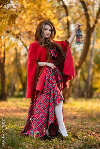 A young woman stands in an autumn forest. She's wearing a long red plaid dress and a warm cape, a romantic, vintage style. She's holding a lantern.
