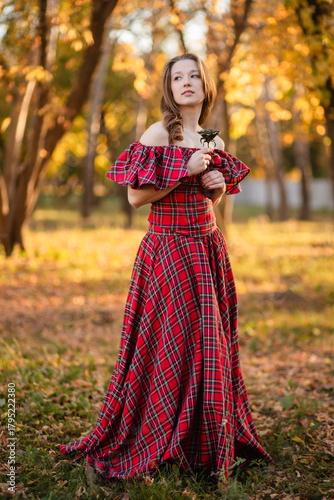 A young woman stands in an autumn forest. She is wearing a long, red, plaid dress with off-the-shoulder details and a frill, a romantic, vintage style. She holds a decorative metal flower in her hands
