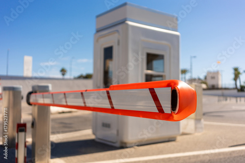 Fototapeta Naklejka Na Ścianę i Meble -  Red and white security barrier gate blocking road entrance near small guard booth under bright sunlight, modern parking access control system outdoors on clear summer day