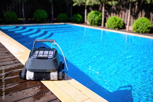 A pool cleaning robot is placed on the edge of an outdoor swimming pool with clear blue water. Green bushes and trees in the background create a cozy private backyard atmosphere