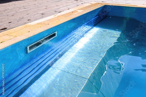 A skimmer installed in the pool wall removes debris from the water surface, maintaining cleanliness. Bright sunlight creates shimmering reflections on the blue water