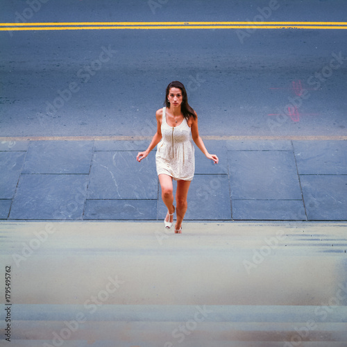 Woman Walking Upstairs on a City Street. A woman in a white dress ascends a set of stairs, with the road and yellow lines visible in the background. Her expression enhances the urban atmosphere.