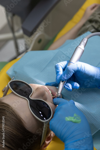 Pediatric dentist in blue gloves works with dental drill near lower teeth while child wears dark protective glasses.