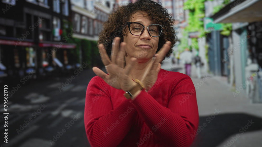 Fototapeta premium Woman gently touches curly hair in red sweater on a bright sunny bustling city street; quiet confidence.