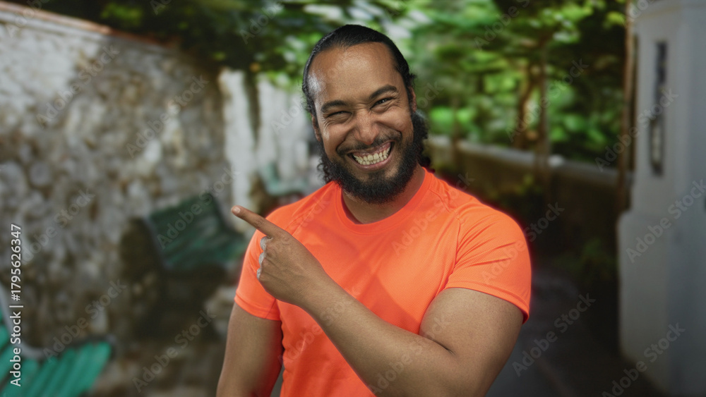 Fototapeta premium Man points finger to the side on a street near a stone wall and bench while wearing a bright orange shirt and ponytail; playful surprise.