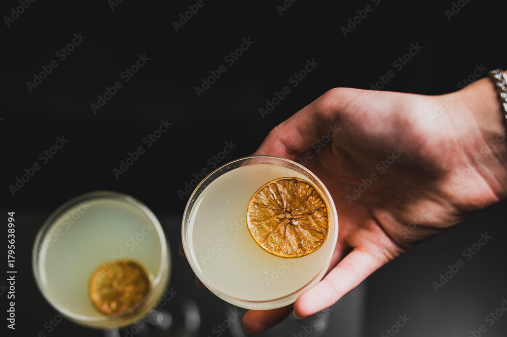 Fototapeta premium A hand holds a yellow cocktail in a coupe glass, garnished with a dried citrus wheel, against a dark background with a second blurred drink nearby