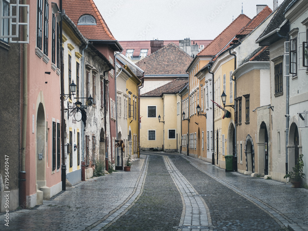 Fototapeta premium Narrow cobblestone street in Sopron, Hungary, lined with colorful historic buildings, arched doorways, and traditional lanterns on a quiet, overcast day.