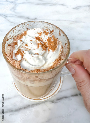 Hand holds cup of hot chocolate or latte coffee with whipped cream and cinnamon