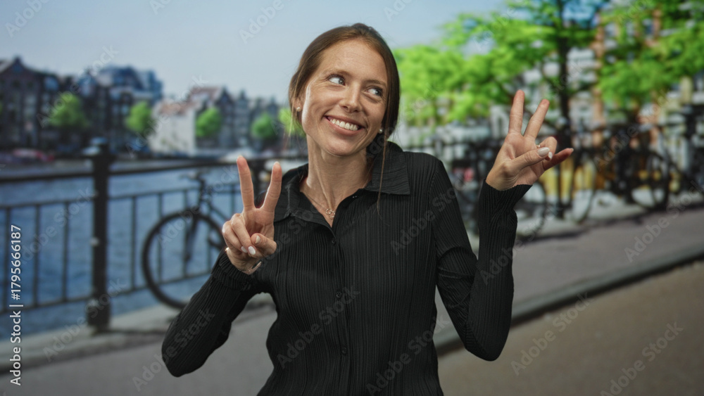 Fototapeta premium Woman holds up peace sign with both hands on amsterdam street by canal bridge, smiling broadly; joy travel fun.