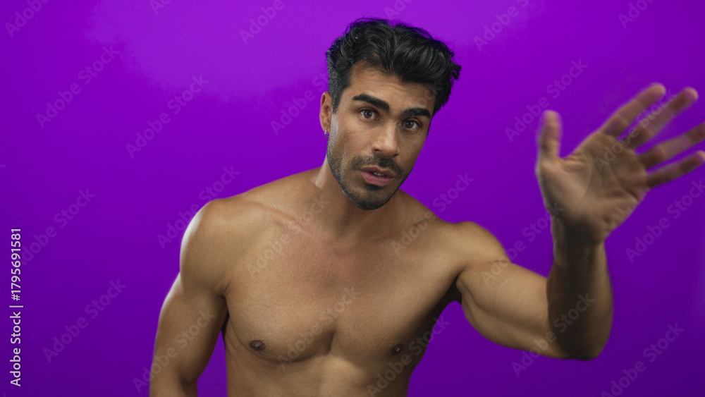 Fototapeta premium Young hispanic man with muscular bare chest points fingers downward in purple studio; invitation.