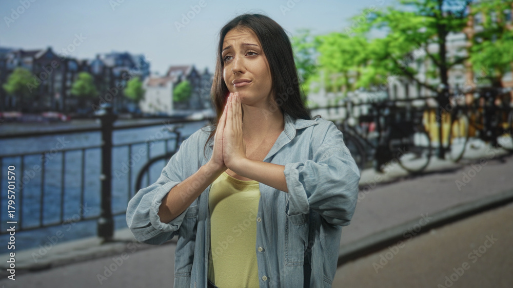 Fototapeta premium Young hispanic woman with hands clasped in prayer and closed eyes on amsterdam street by canal railing, wearing denim shirt; hope plea.