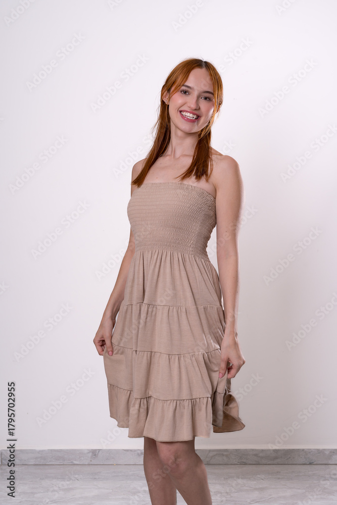Fototapeta premium Red-haired woman smiling with calm and confident expression, beige dress studio fashion portrait.